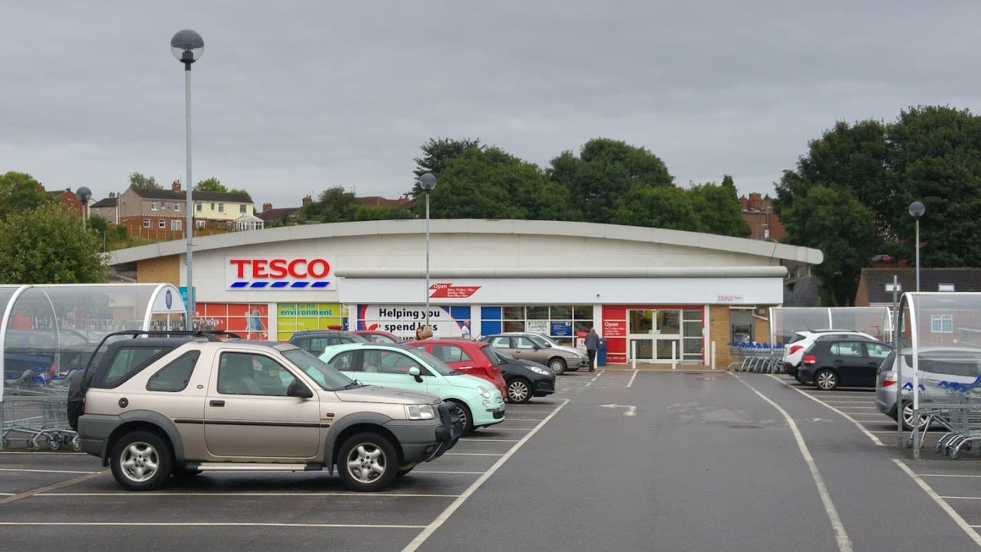 Tesco Superstore entrance with parking lot, shopping carts, and trees in the background under a cloudy sky.