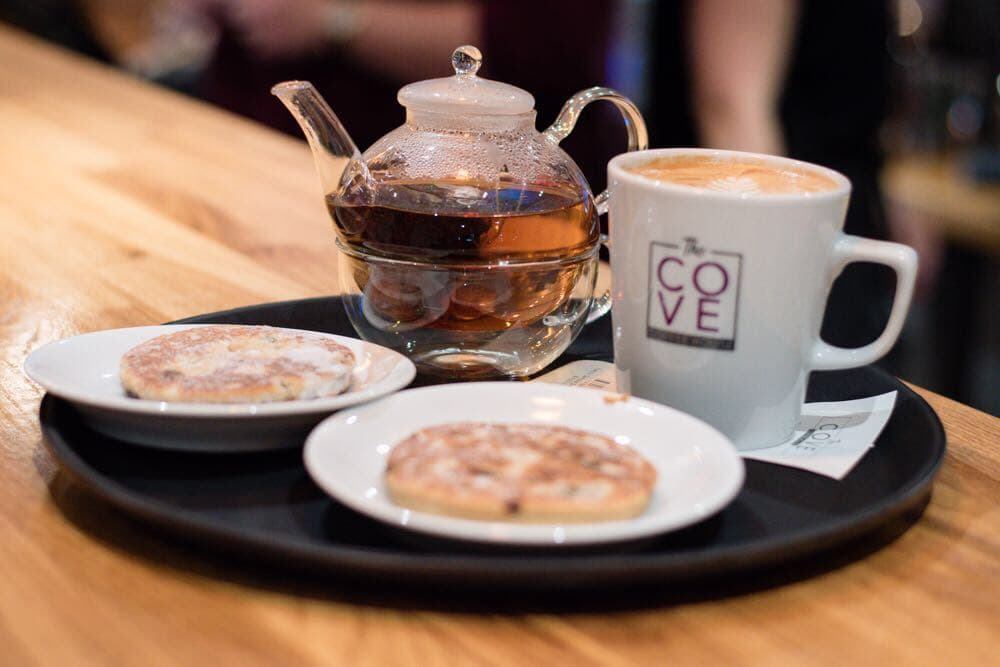 A tray with two cookies, a teapot, and a coffee cup bearing "The Cove" logo, set on a wooden counter.