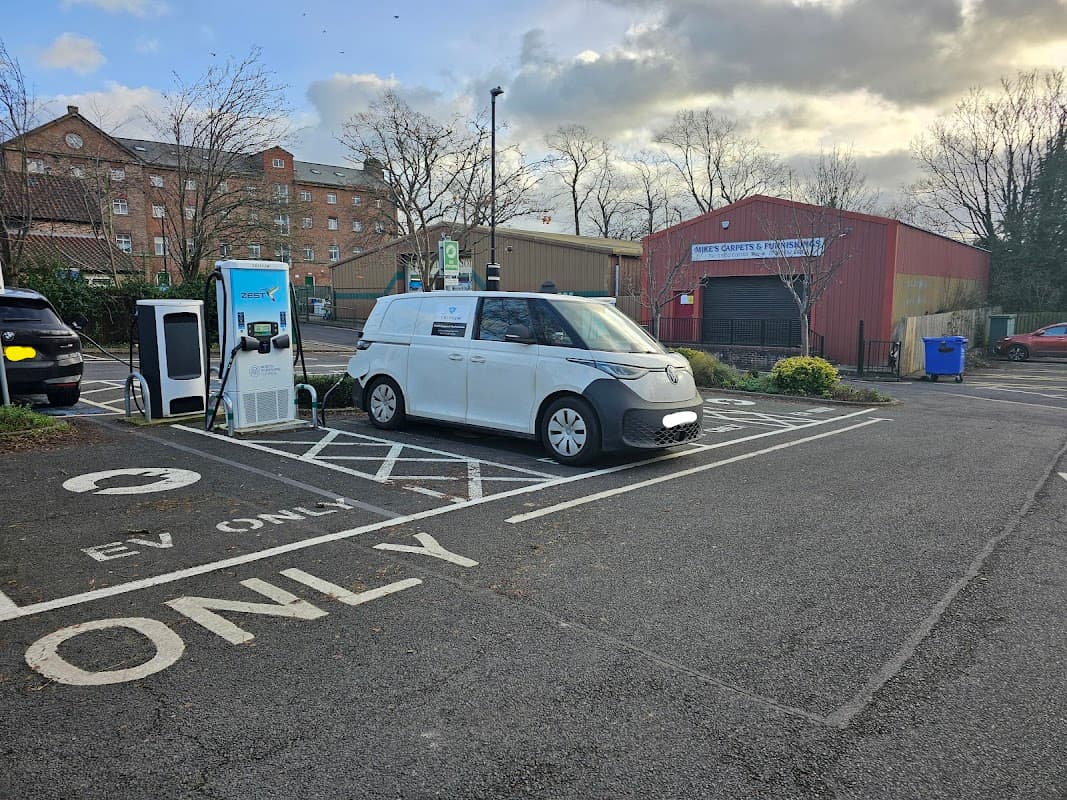 Electric vehicle charging station with a parked van in a car park, surrounded by trees and buildings.