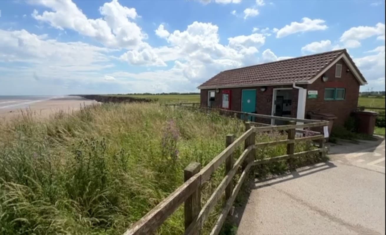Pay & Display parking area near Mappleton Beach, with a small building and grassy surroundings under a blue sky.