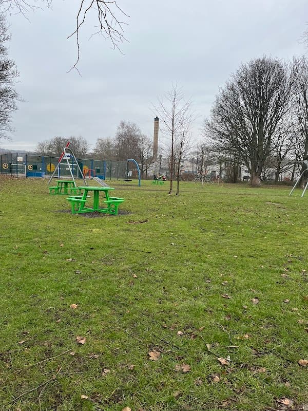 Green picnic tables on grass with playground equipment and trees in Darton Park, Mapplewell, Yorkshire.