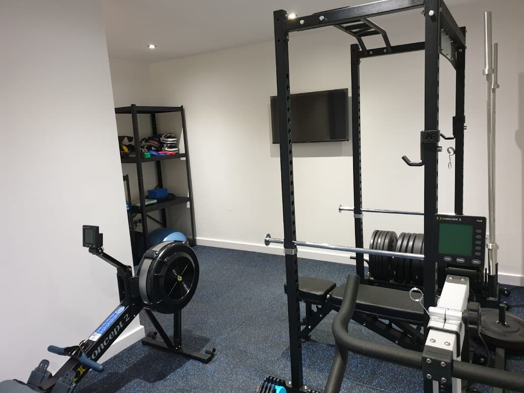 Fitness equipment including a rowing machine, weight rack, and exercise bench in a well-lit gym space.