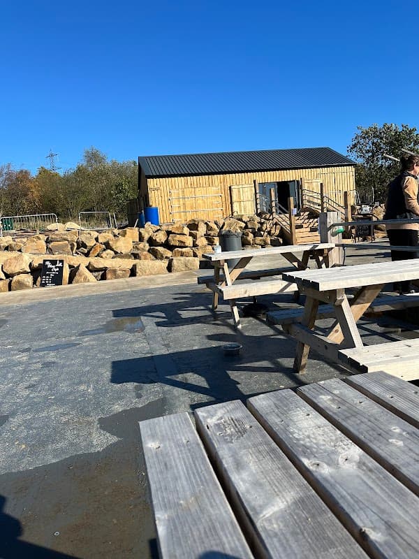Wooden farm building with a black roof, surrounded by large rocks and picnic tables under a clear blue sky.