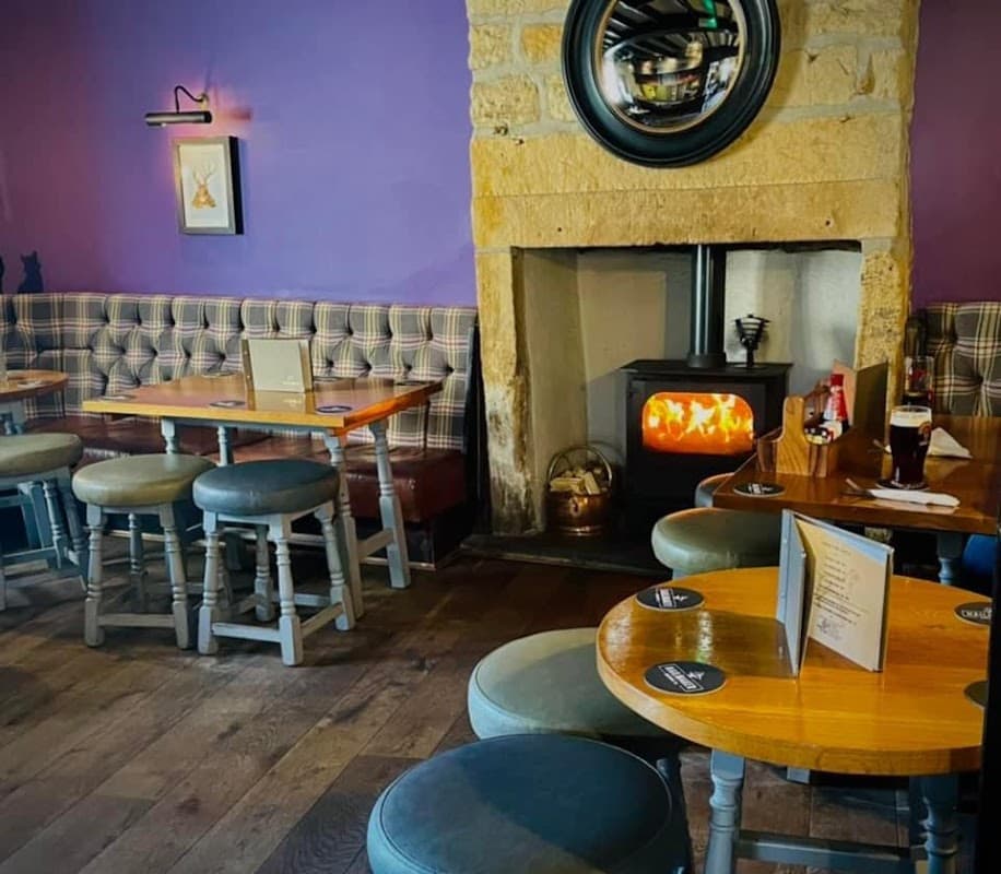 Cozy bar interior with stone fireplace, wooden tables, plaid seating, and warm lighting in Mapplewell, Yorkshire.