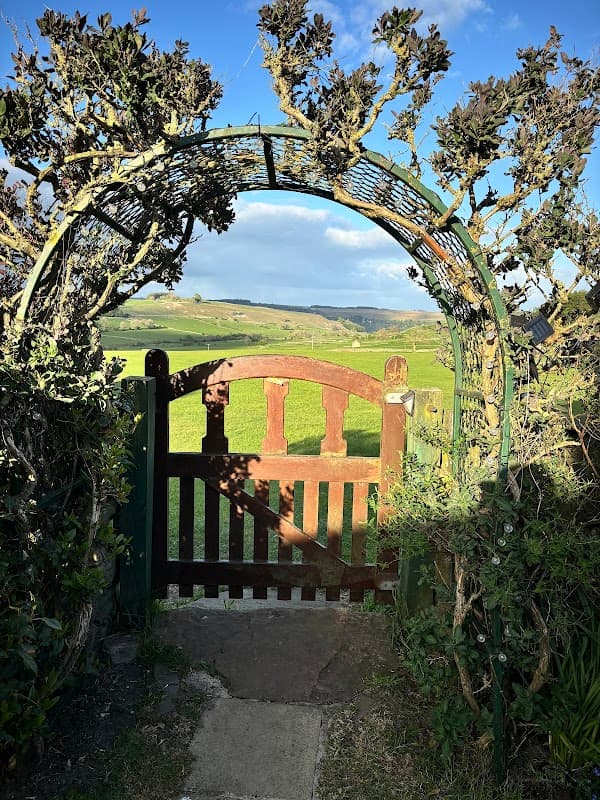 Rustic wooden gate framed by greenery, opening to lush green fields and rolling hills under a clear blue sky.