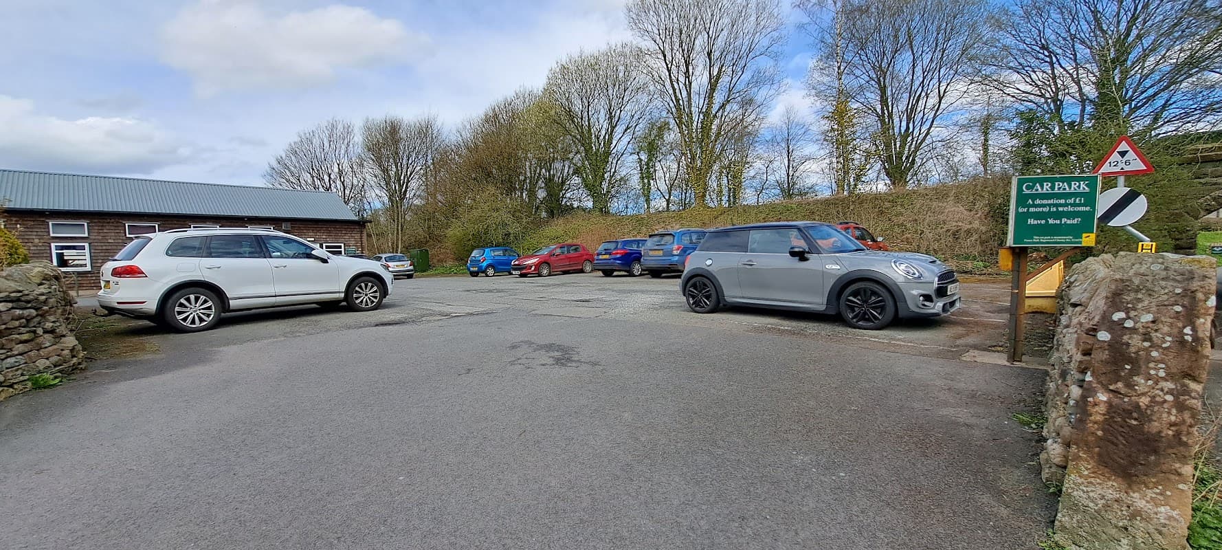 Cowan Bridge Carpark with several parked cars, stone walls, and trees under a partly cloudy sky.