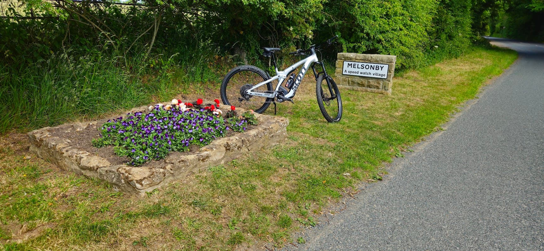 A bicycle rests beside a flower bed with colorful blooms and a sign reading "MELSONBY, a good place to live."