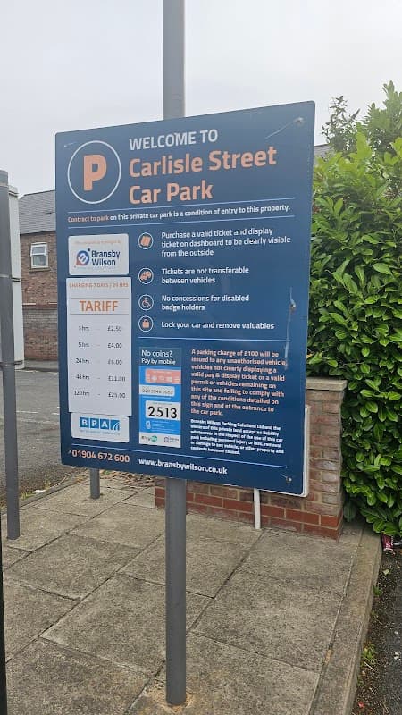 Welcome sign for Carlisle Street Car Park with parking rates and instructions, surrounded by greenery and buildings.