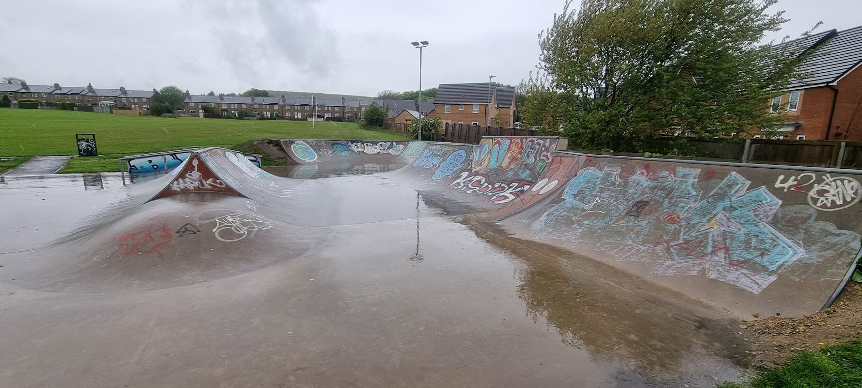 Skatepark with wet concrete ramps, colorful graffiti, and grass fields in the background under a cloudy sky.