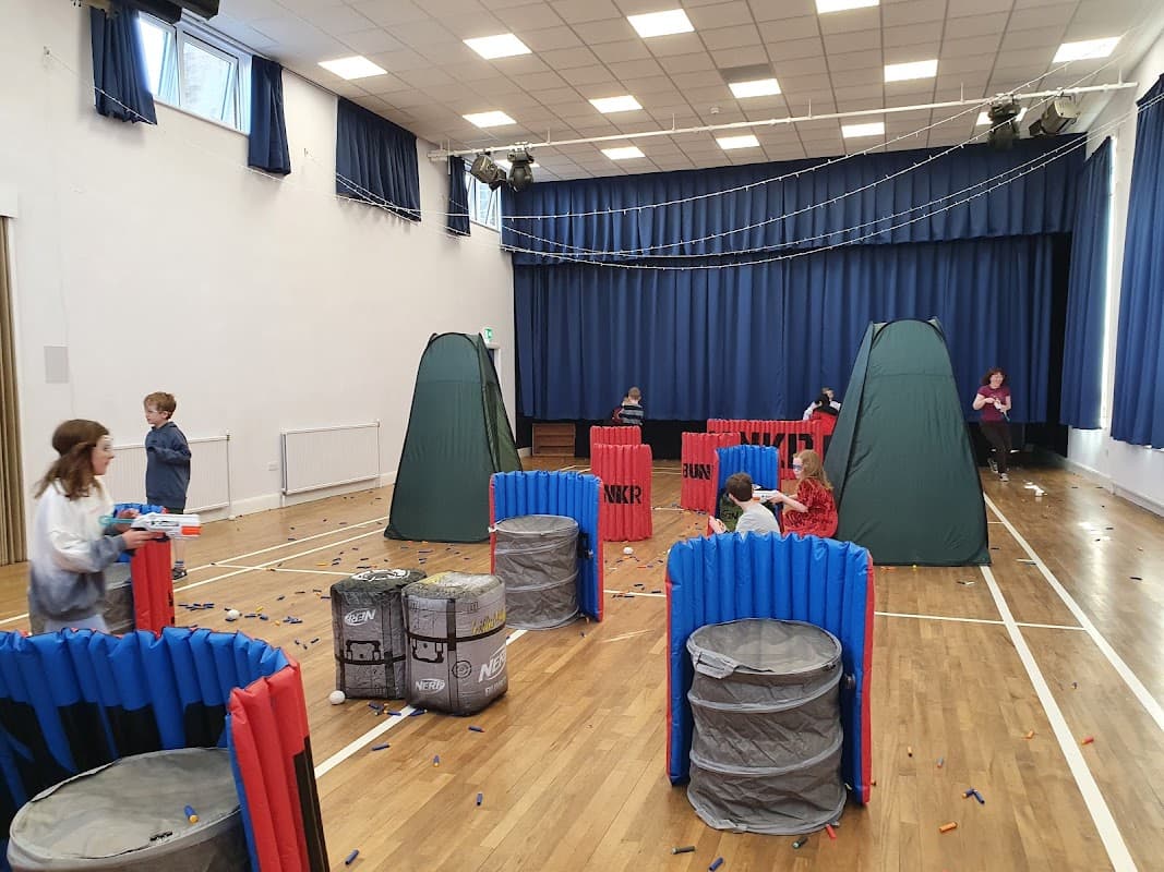 Community center hall with colorful barriers, tents, and children playing in a Nerf gun battle setup.