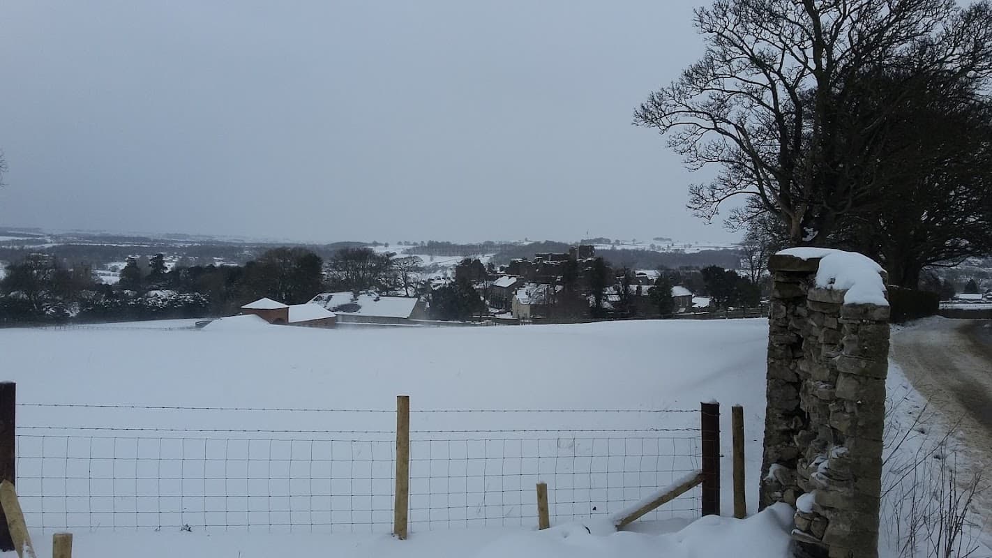 Snow-covered landscape with a stone wall, fence, and distant buildings under a grey sky in Middleham, Yorkshire.