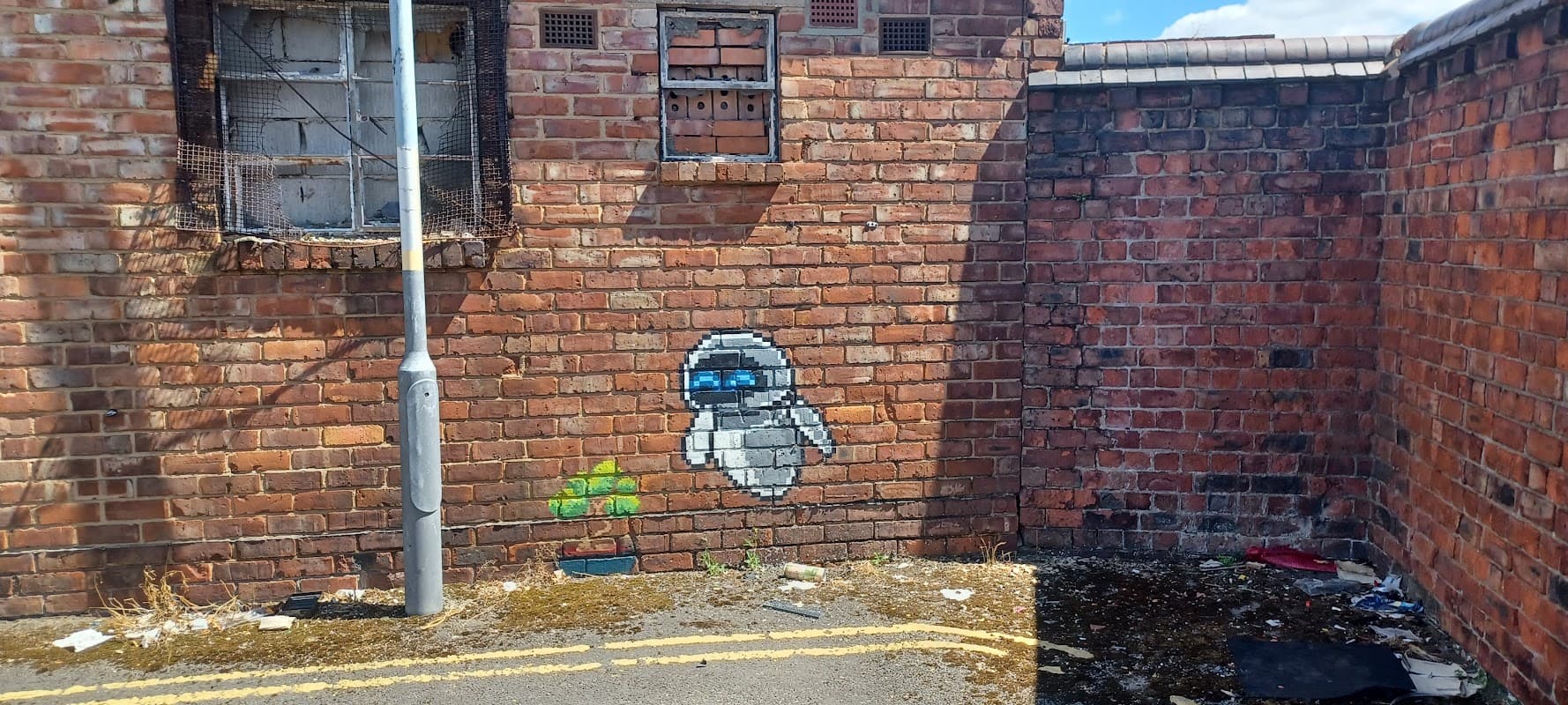 Graffiti of a ghost-like figure on a brick wall, surrounded by litter and overgrown weeds in a car park.