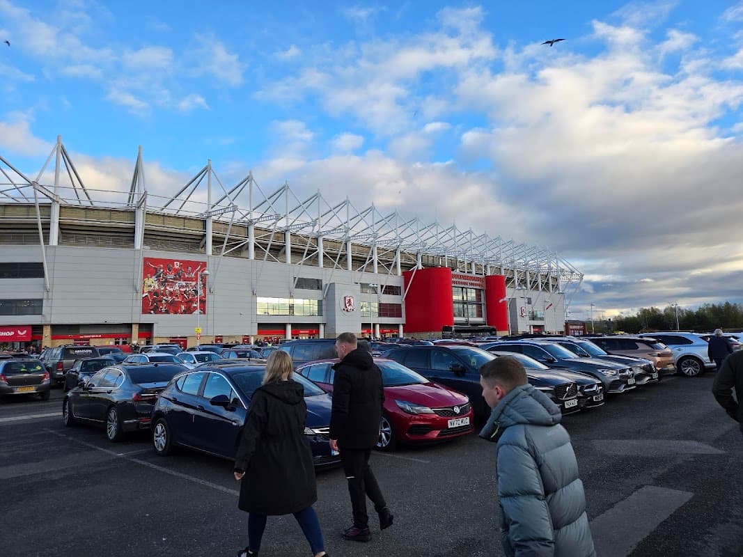 Busy car park with numerous parked cars and people walking, alongside a large stadium under a cloudy sky.