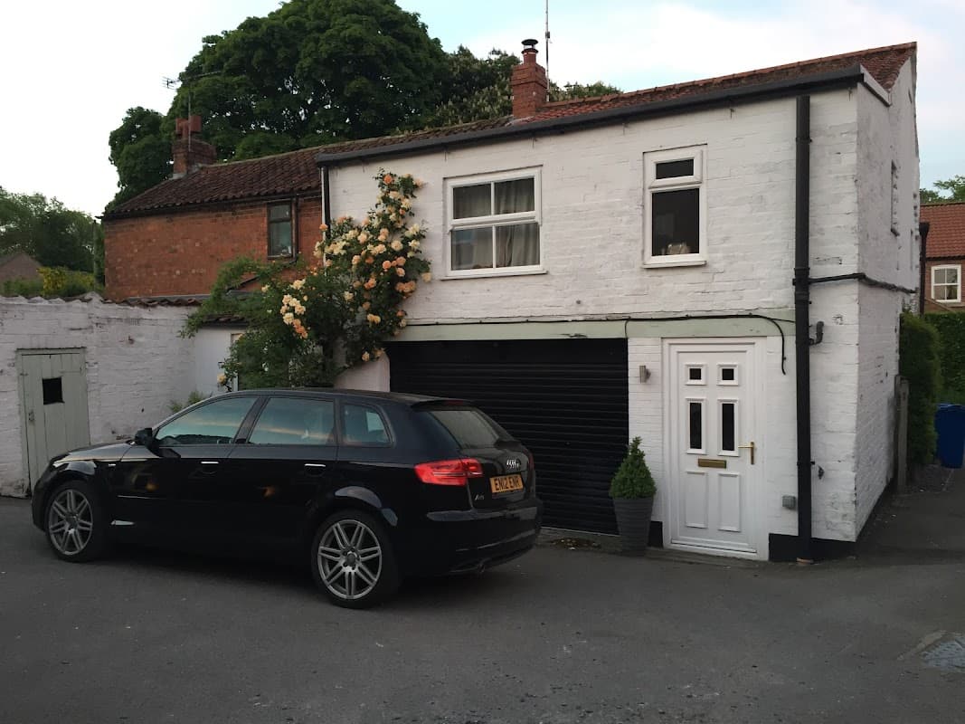 A white two-story building with a black car parked in front, surrounded by greenery and a rose bush.