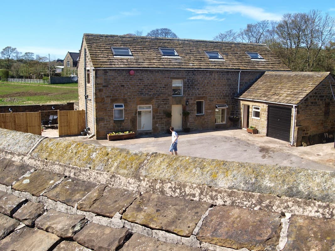 Stone barn with a slate roof, surrounded by greenery, featuring a driveway and a person walking nearby.