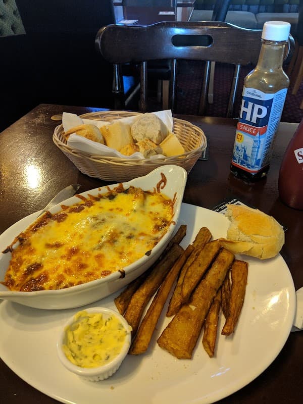 A plate of cheesy lasagna with fries, a roll, and a basket of bread beside a bottle of HP sauce on a wooden table.