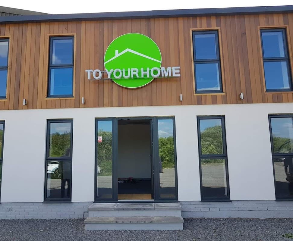 Modern shop exterior with large green sign "TO YOUR HOME" and large windows, located in Millhouse Green, Yorkshire.