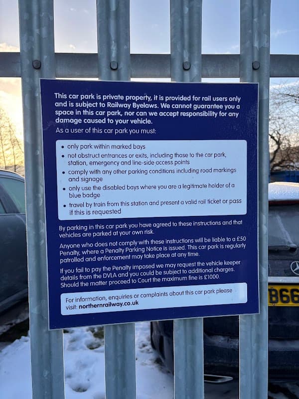 Sign detailing parking rules and regulations at Mirfield Station Car Park, with a fence and vehicles visible in the background.