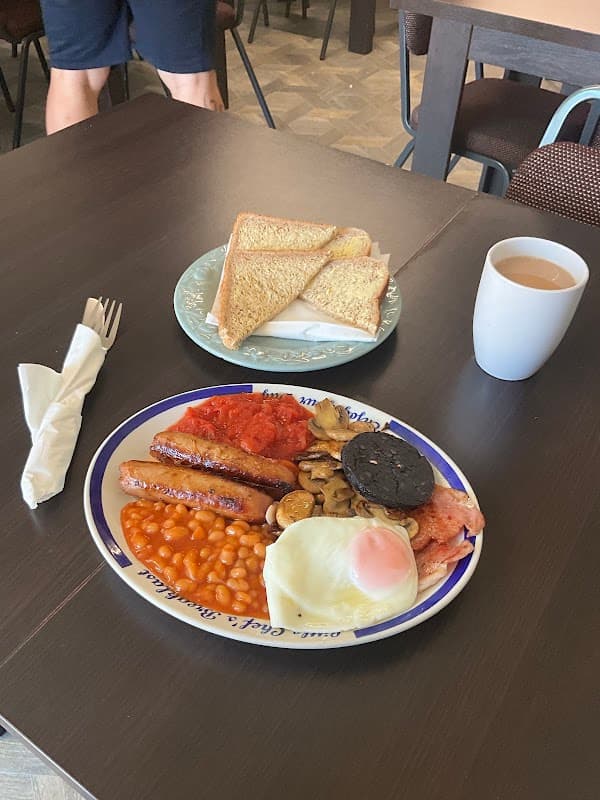 A full English breakfast with sausages, beans, mushrooms, bacon, a fried egg, toast, and a cup of tea on a table.