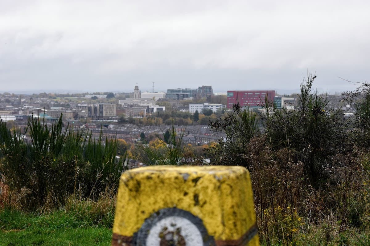 Yellow trig point in foreground with a panoramic view of a town and cloudy sky in the background.