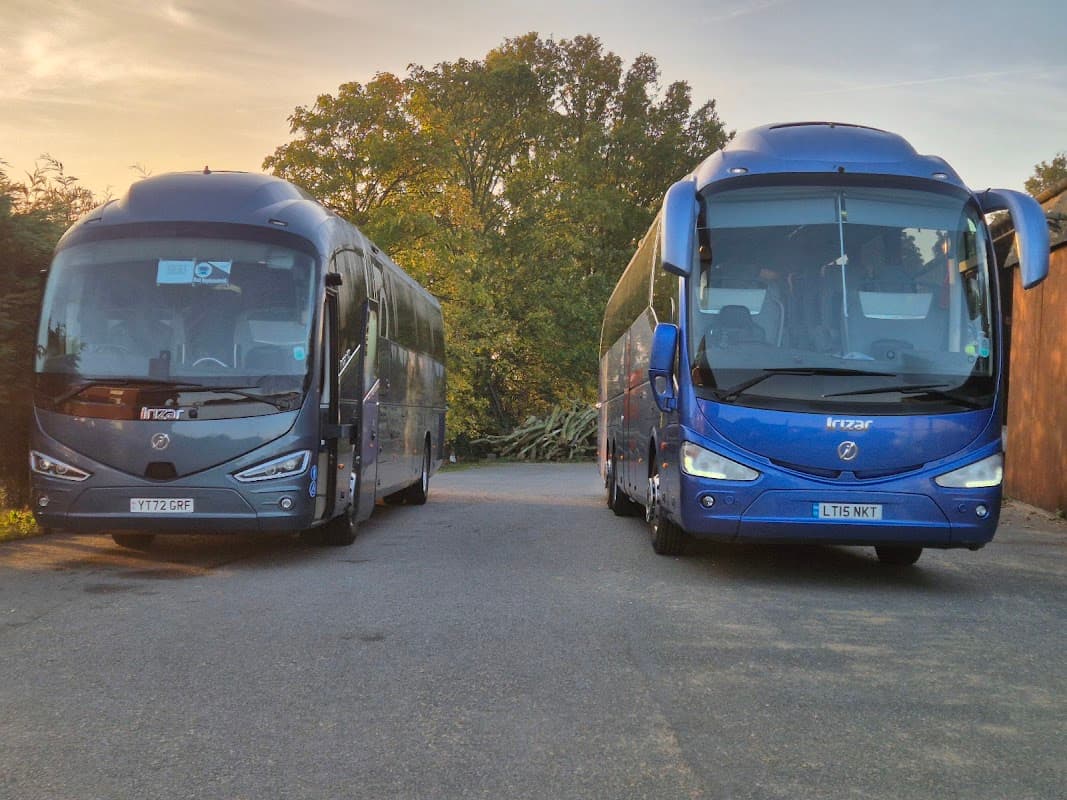 Two blue coaches parked side by side on a road, surrounded by trees in the background, during sunset.