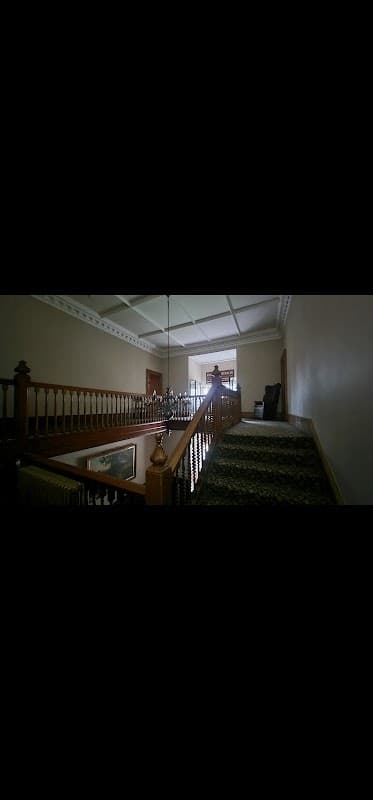 Staircase with wooden railing and carpet, leading to a well-lit room with large windows in a historic building.