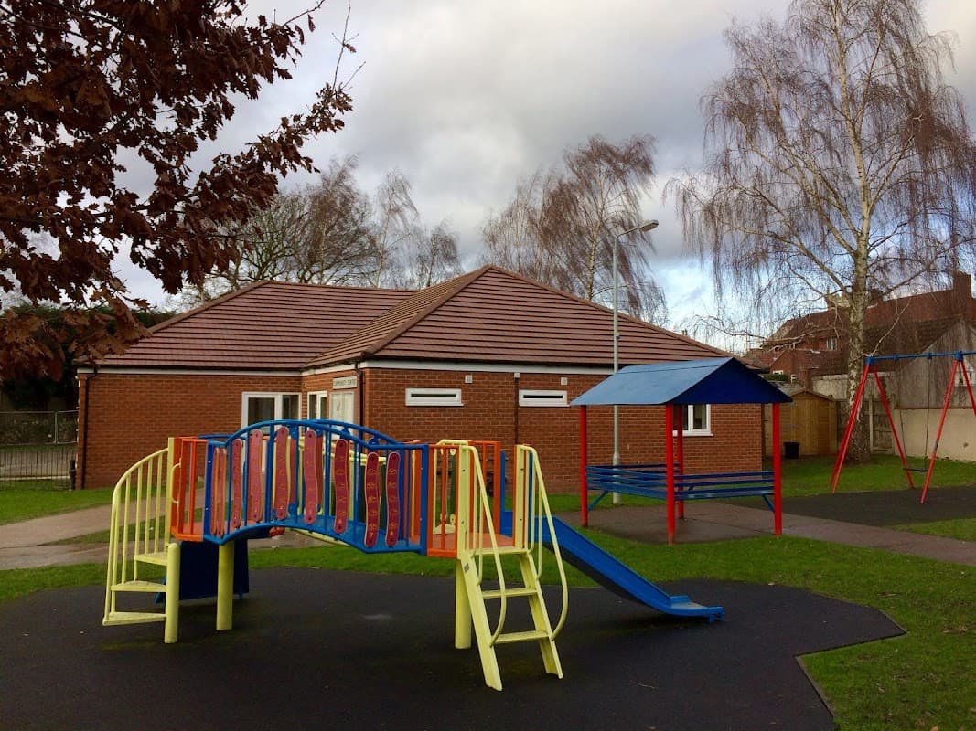 Community centre with a playground featuring a slide, bridge, and swings, surrounded by trees and cloudy skies.