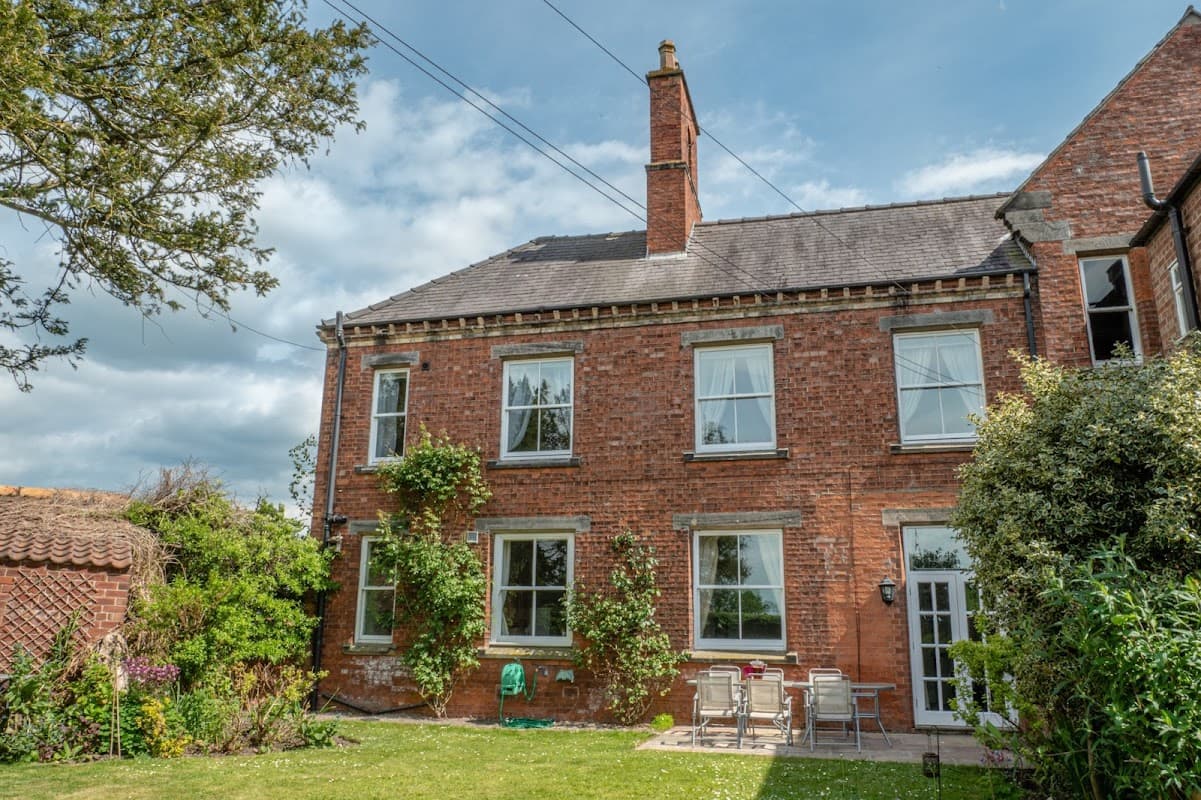 Red brick house with large windows, green garden, patio furniture, and surrounding trees under a partly cloudy sky.