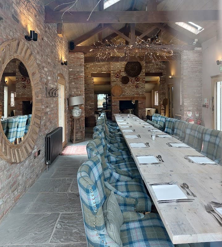 Long wooden dining table with blue plaid seating, exposed brick walls, and decorative lighting in a cozy restaurant interior.