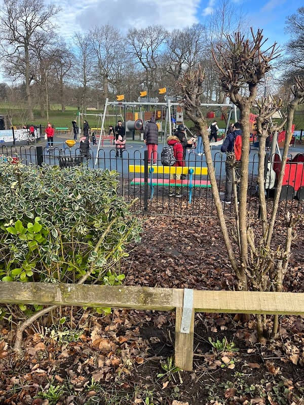 Lakeside park with a playground, children playing, and trees in a grassy area, surrounded by a black fence.