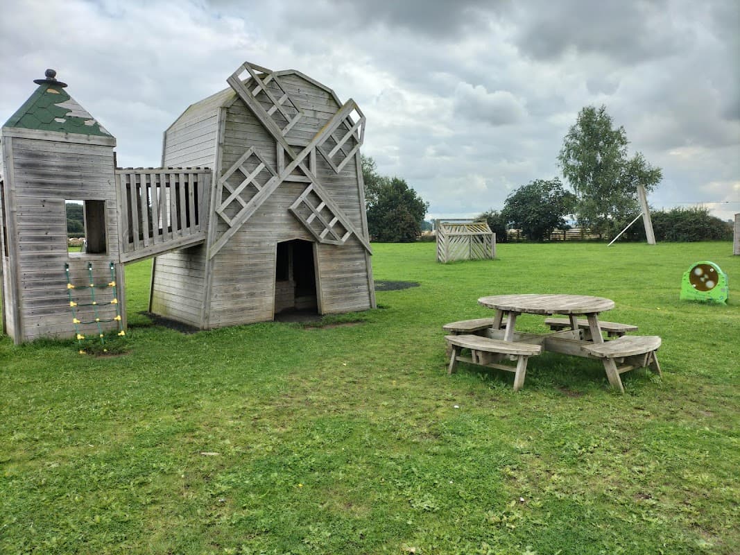 Wooden playground structure resembling a windmill, picnic table, and green grass under a cloudy sky.