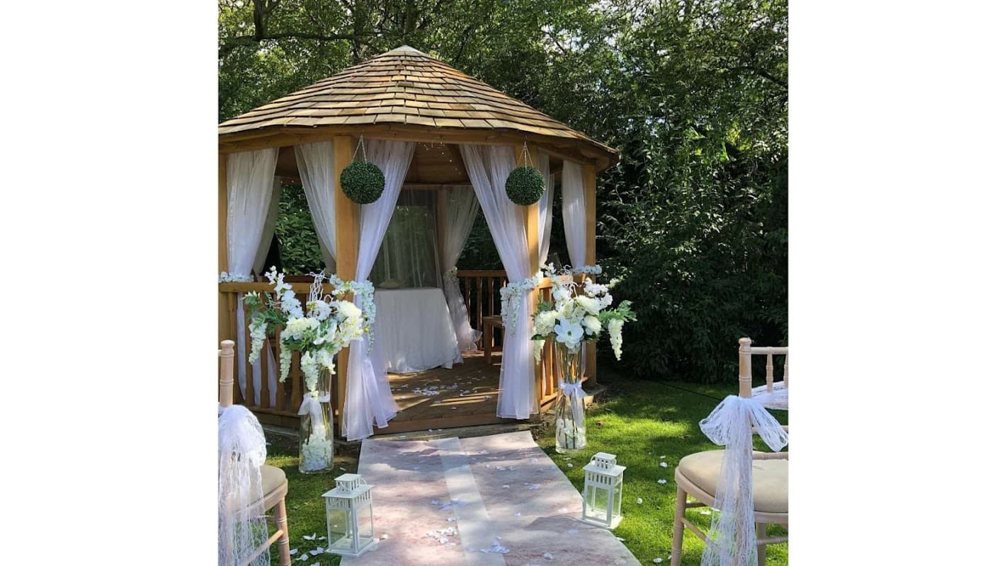 A charming gazebo adorned with flowers and drapes, surrounded by greenery, set for a wedding ceremony.