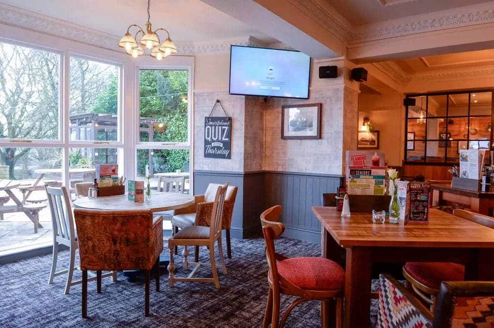 Cozy bar interior with wooden tables, chairs, a TV, and a quiz sign, overlooking a garden area through large windows.