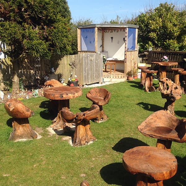 Wooden furniture in a grassy area, with carved tables and chairs, surrounded by greenery and a small shed.