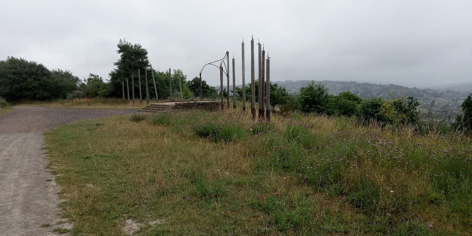 Grass-covered path leading to tall metal sculptures amidst greenery and a misty landscape.