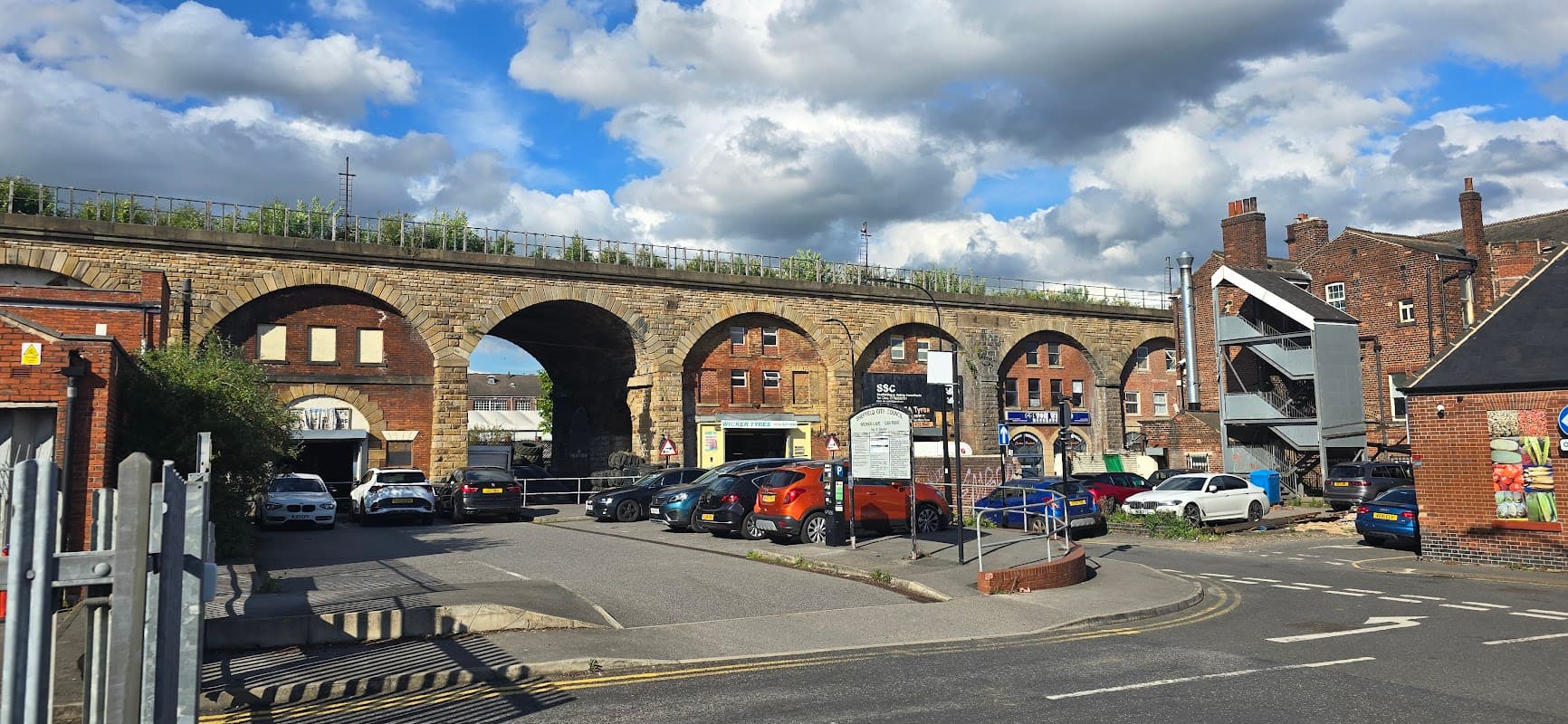 Wicker Lane Car Park with parked cars, a stone viaduct above, and brick buildings under a cloudy sky.