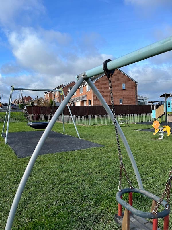 Playground with swings, a spring rider, and grassy areas, surrounded by houses under a blue sky with clouds.