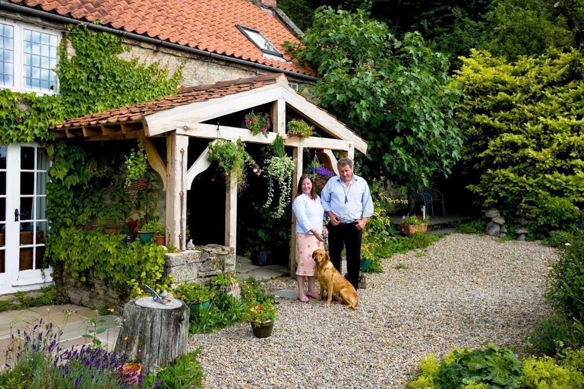 Charming bed and breakfast with a wooden entrance, lush greenery, a couple, and a dog in a gravel courtyard.