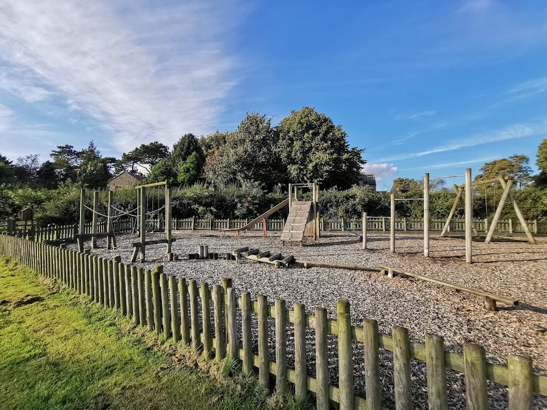 Play park with climbing frames, slides, and a gravel surface, surrounded by wooden fencing and greenery under a blue sky.