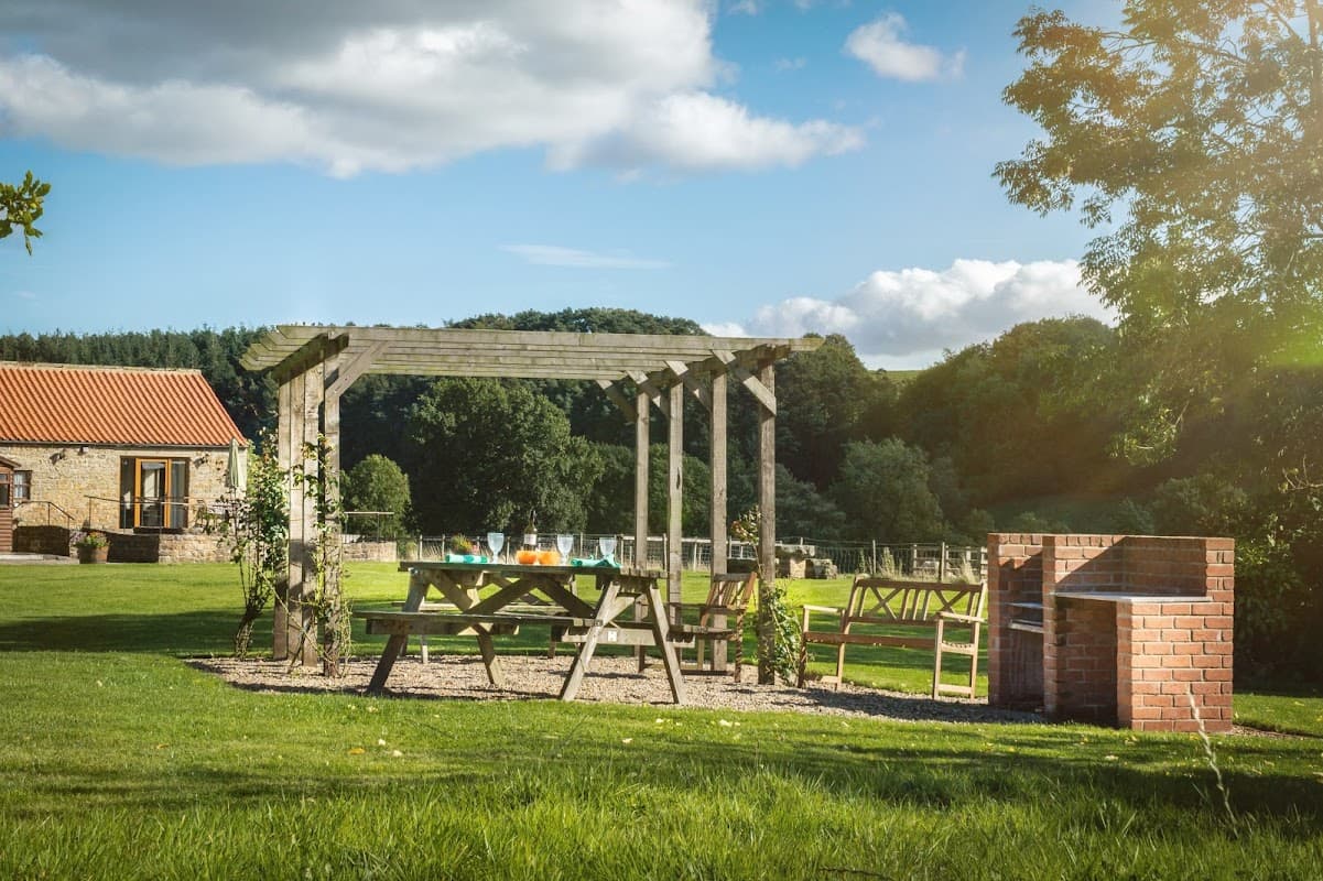 Rustic wooden pergola with picnic table and chairs in a grassy area, surrounded by trees and a brick barbecue.