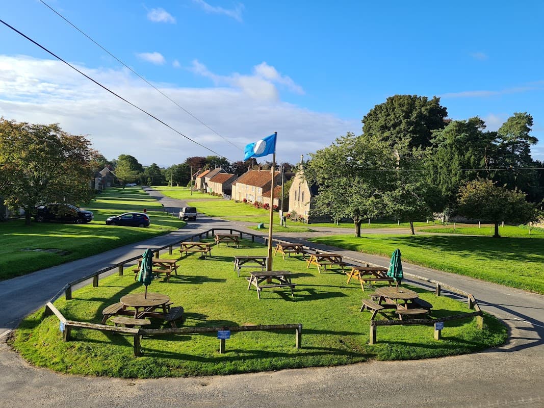 The Horseshoe Inn features a grassy area with picnic tables, a flagpole, and surrounding trees in a quaint village setting.