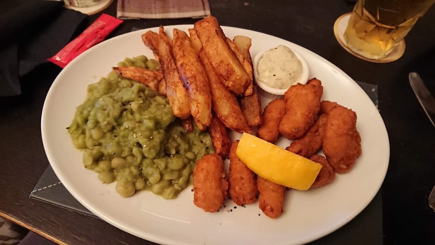 Plate of golden-brown battered fish, chips, mushy peas, a lemon wedge, and a small bowl of tartar sauce.