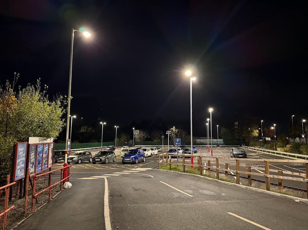 Well-lit car park at night with several parked cars, trees, and a red fence in the foreground.
