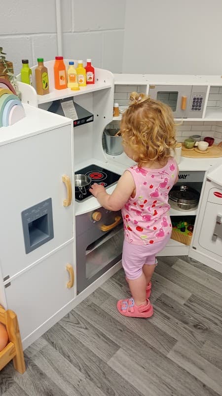 A toddler with curly hair plays at a toy kitchen, pretending to cook with various colorful food items around.