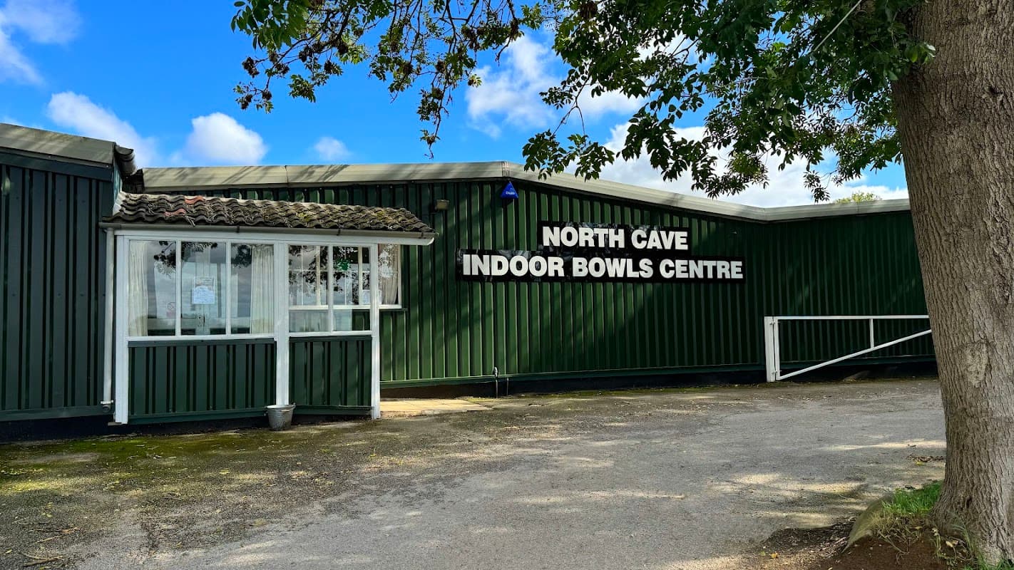 North Cave Indoor Bowls Centre sign on a green building with a glass entrance and tree nearby. Blue sky and clouds above.
