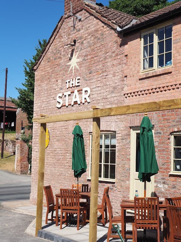 Brick building with "THE STAR" sign, green umbrellas, and wooden tables outside in a sunny setting.