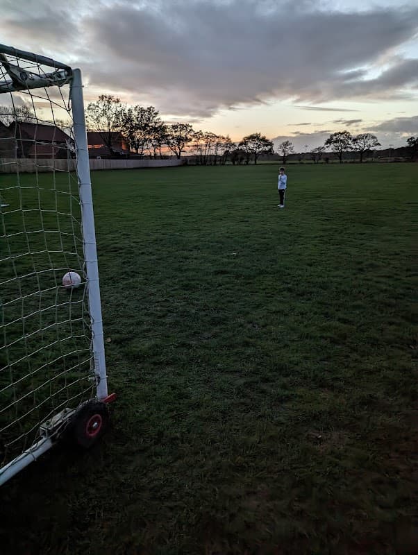 A child stands in a football field near a goalpost at sunset, with trees silhouetted in the background.