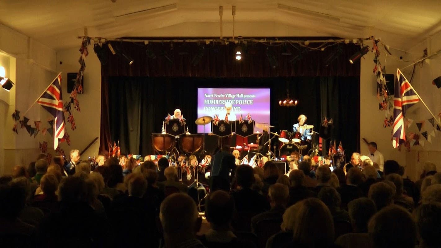 North Ferriby Village Hall filled with an audience, featuring musicians, British flags, and a stage backdrop.