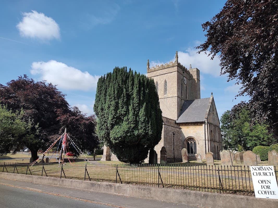 Norman church with tall trees, gravestones, and a sign reading "Open for Coffee" against a blue sky.