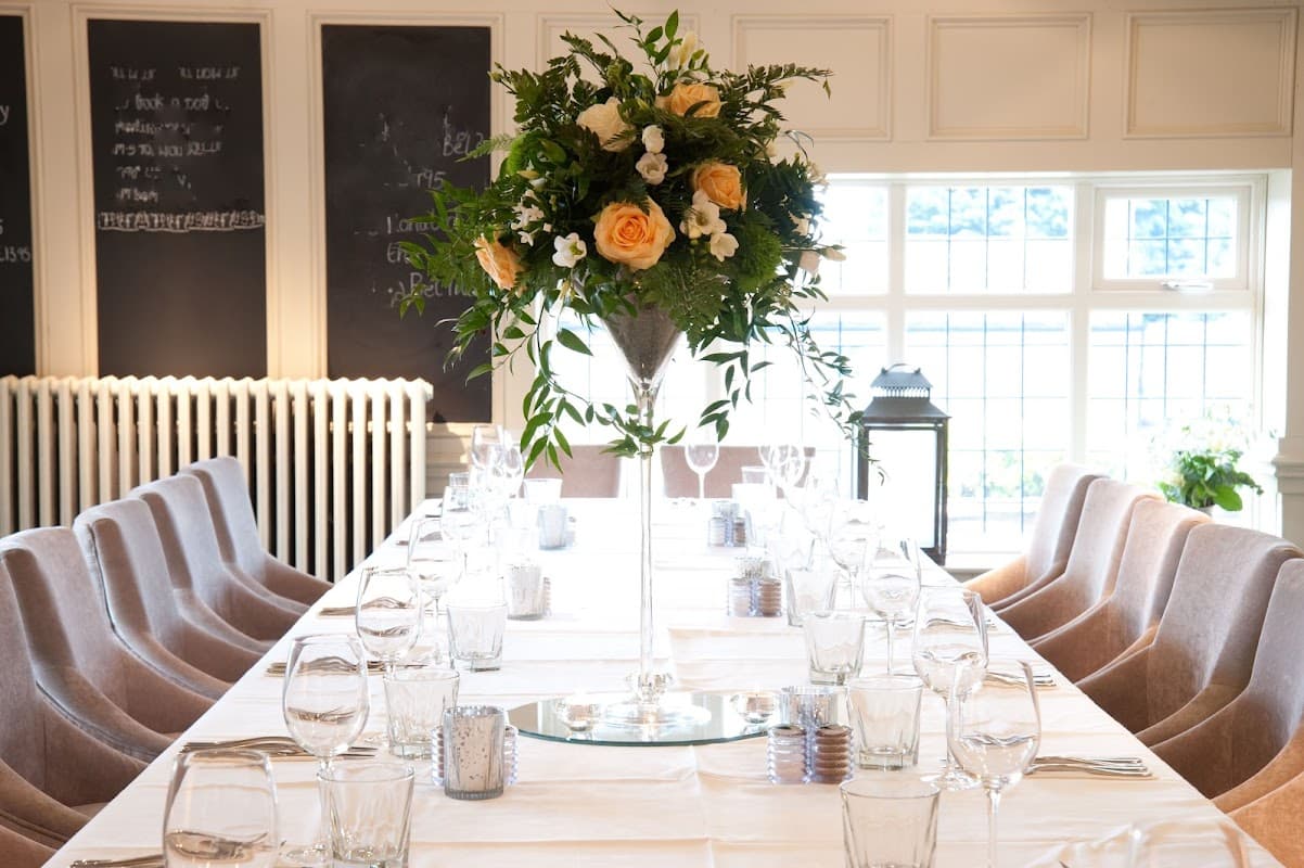 Elegant dining setup with a long table, glassware, and a tall floral centerpiece featuring roses and greenery.